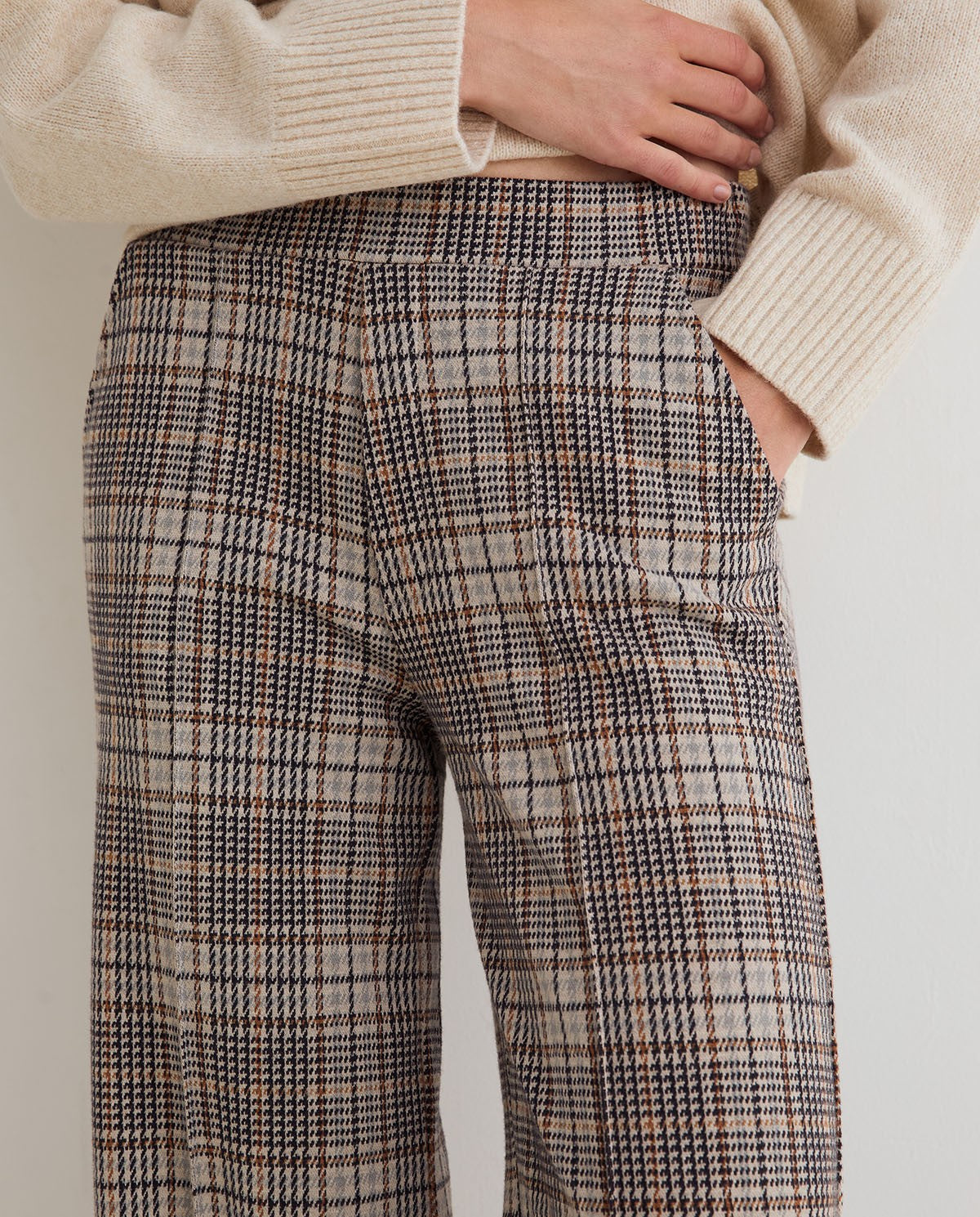 Close-up of a person wearing high-waisted plaid trousers in beige, brown, and black, paired with a cream ribbed sweater.