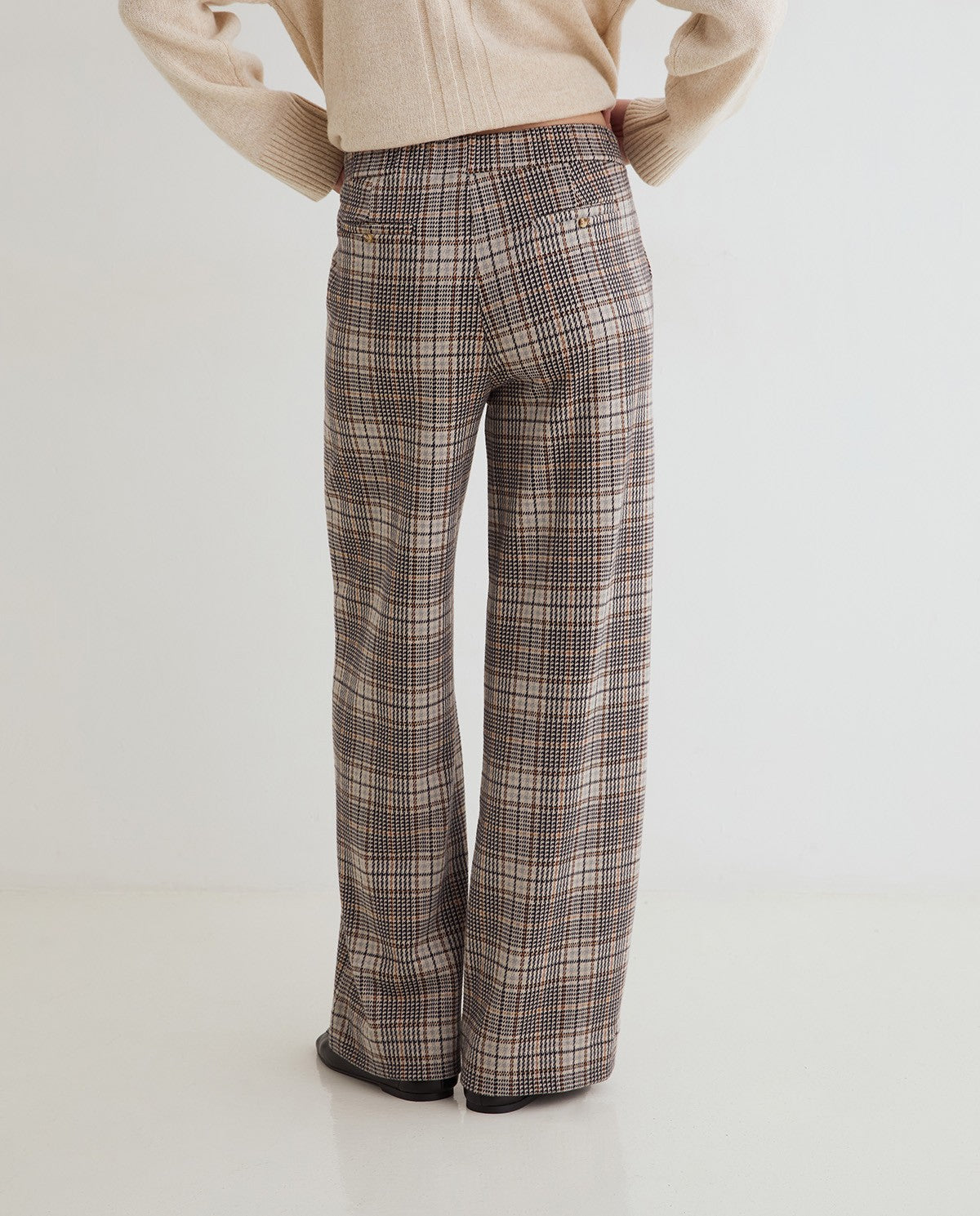 A person shown from the back wearing high-waisted, wide-leg plaid trousers in a brown, beige, and gray pattern, paired with a cream knit sweater.