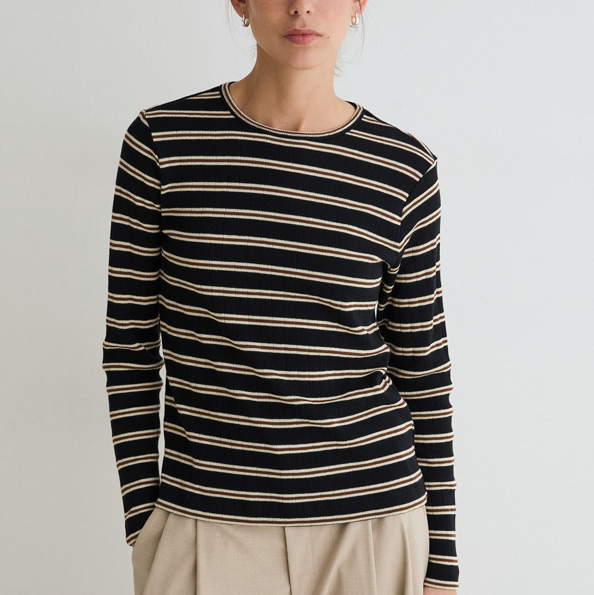 A woman wearing a long-sleeve crewneck knit top with horizontal black and beige stripes, paired with light beige trousers, photographed against a plain light gray studio background.