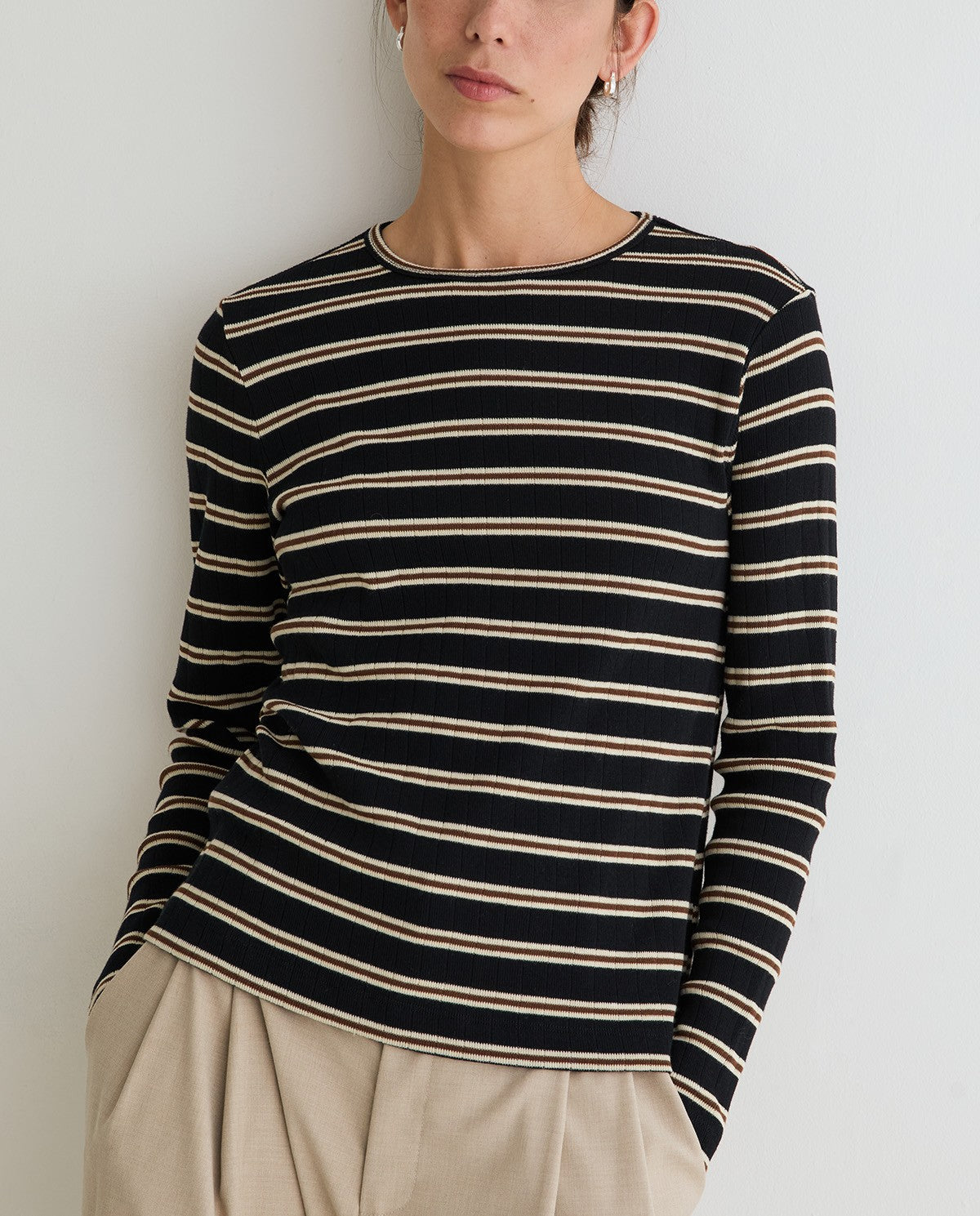 A woman wearing a black long-sleeve knit top with thin beige horizontal stripes, paired with beige trousers, photographed against a plain light gray wall.