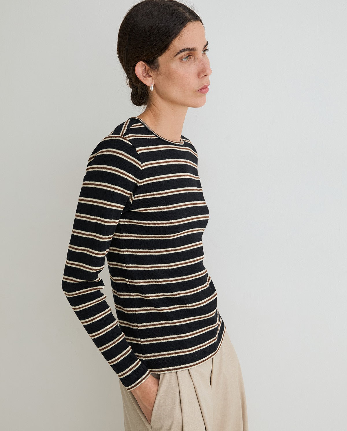 A woman wearing a black long-sleeve knit top with thin beige horizontal stripes, paired with light beige trousers, photographed against a plain light gray studio background.