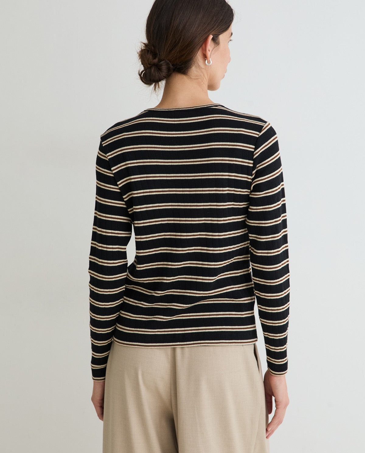 A woman shown from the back wearing a black long-sleeve knit top with thin beige horizontal stripes and light beige trousers, against a plain light gray studio background.