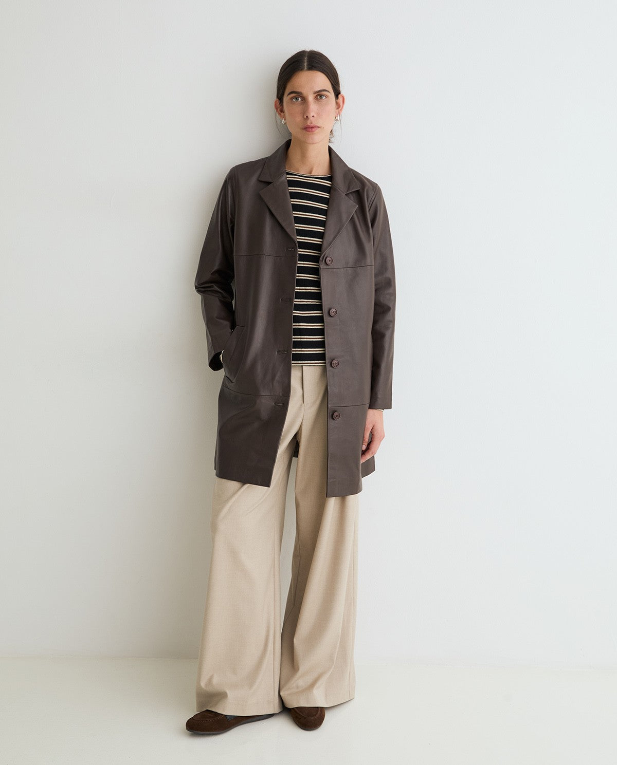 A model stands against a plain white studio wall wearing a dark brown mid-length coat over a black and white striped top, paired with beige wide-leg trousers and dark brown shoes.