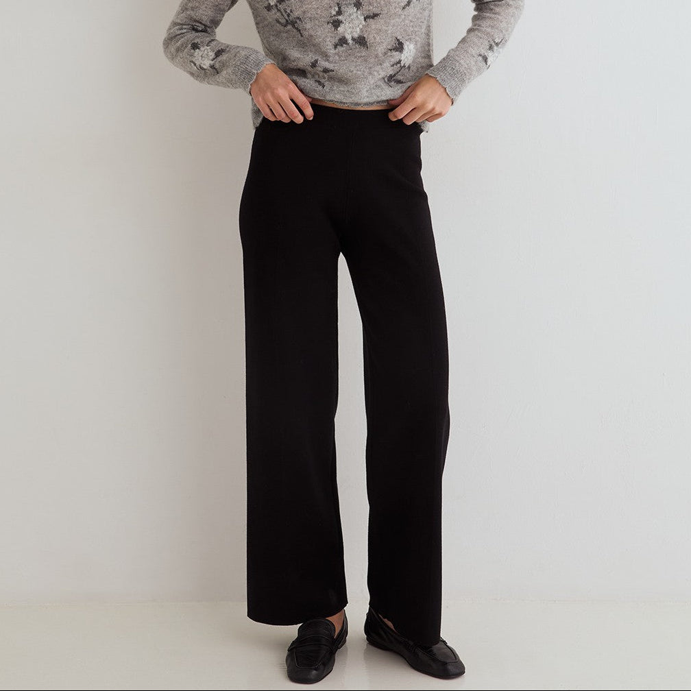 A woman stands against a plain white studio wall wearing a light gray knit sweater with a darker gray floral pattern, black wide-leg trousers, and black loafers.
