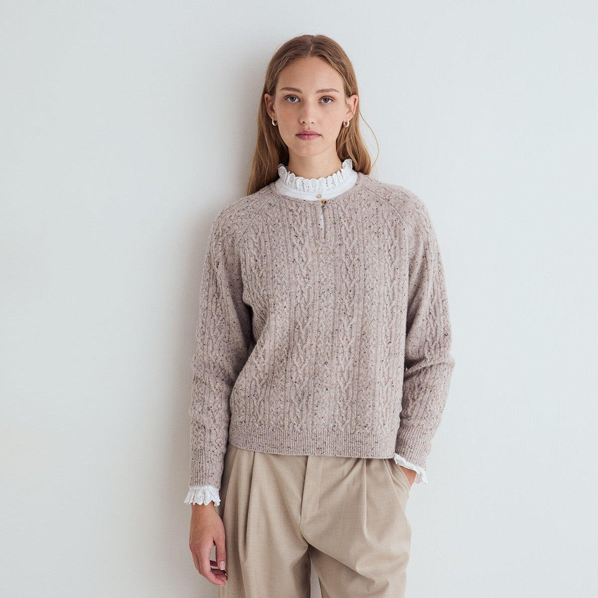 A woman stands against a plain light gray wall wearing a taupe cable-knit sweater over a white ruffled mock-neck top, paired with beige trousers.