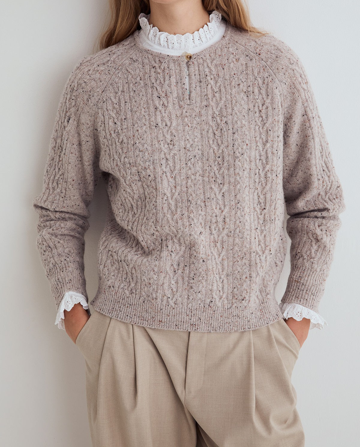 A person wearing a textured cable-knit sweater in a light taupe-beige color, styled with a white ruffled mock neck and cream trousers, against a plain light gray wall.