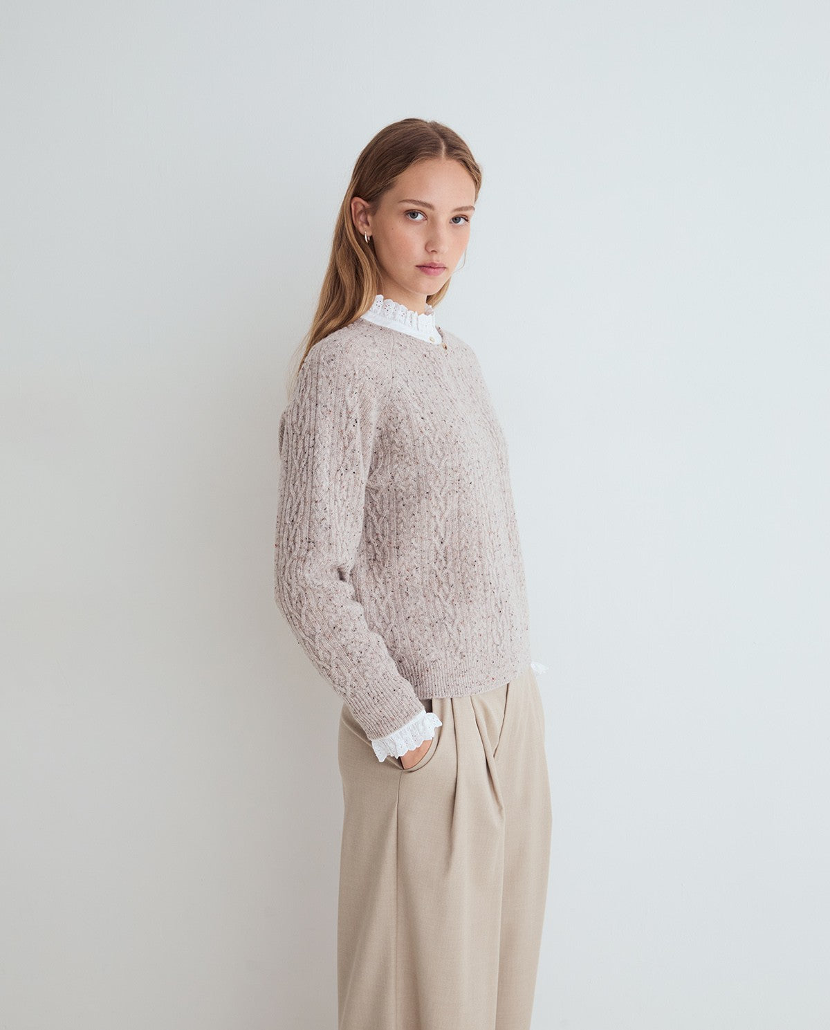 A person wearing a textured cable-knit sweater in a light taupe-beige color, styled with a white ruffled mock neck and cream trousers, against a plain light gray wall.