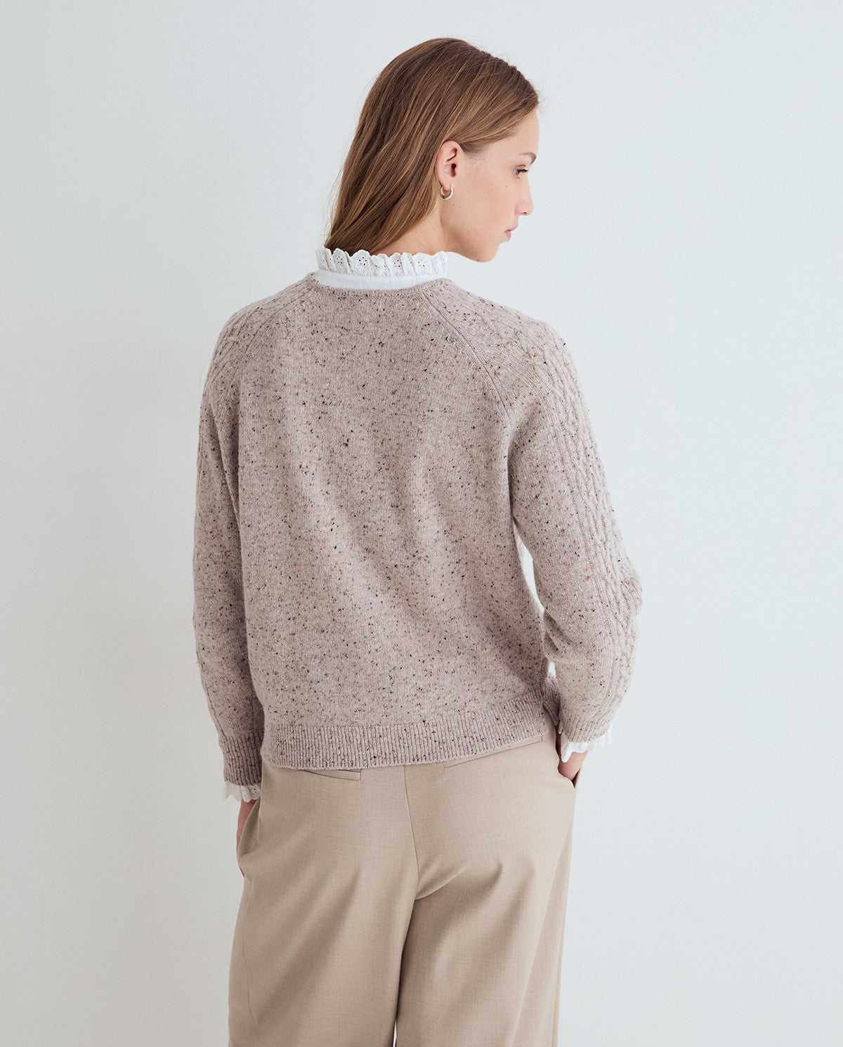 A person wearing a textured cable-knit sweater in a light taupe-beige color, styled with a white ruffled mock neck and cream trousers, against a plain light gray wall.