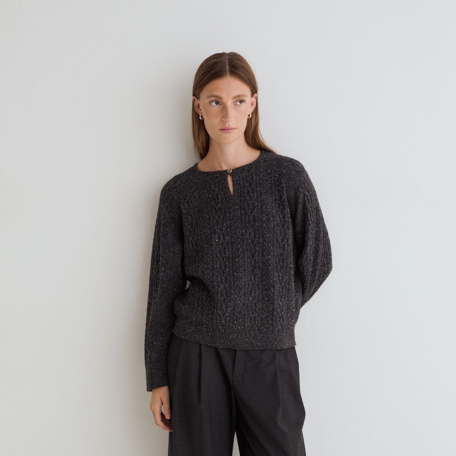 A woman stands against a plain white studio wall wearing a dark charcoal textured knit long-sleeve top with a keyhole neckline, black wide-leg trousers, and red pointed shoes.