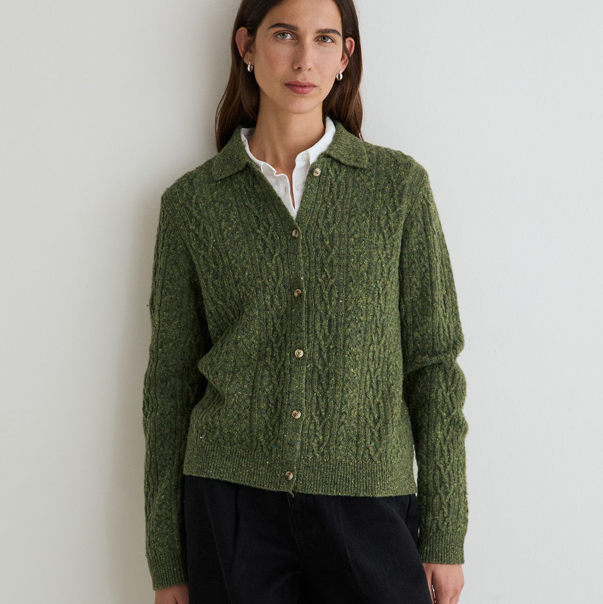 A woman wearing a textured olive green cable-knit button-up cardigan over a white collared shirt and black trousers, standing against a plain light gray wall.