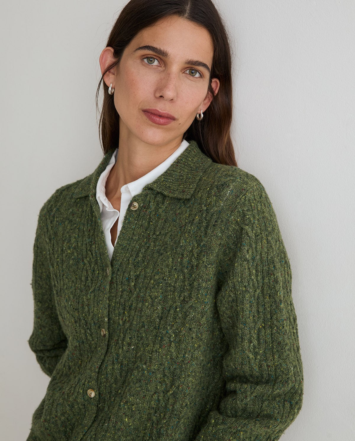 A woman wearing a textured olive green cable-knit cardigan over a white collared shirt, photographed against a plain light gray background.