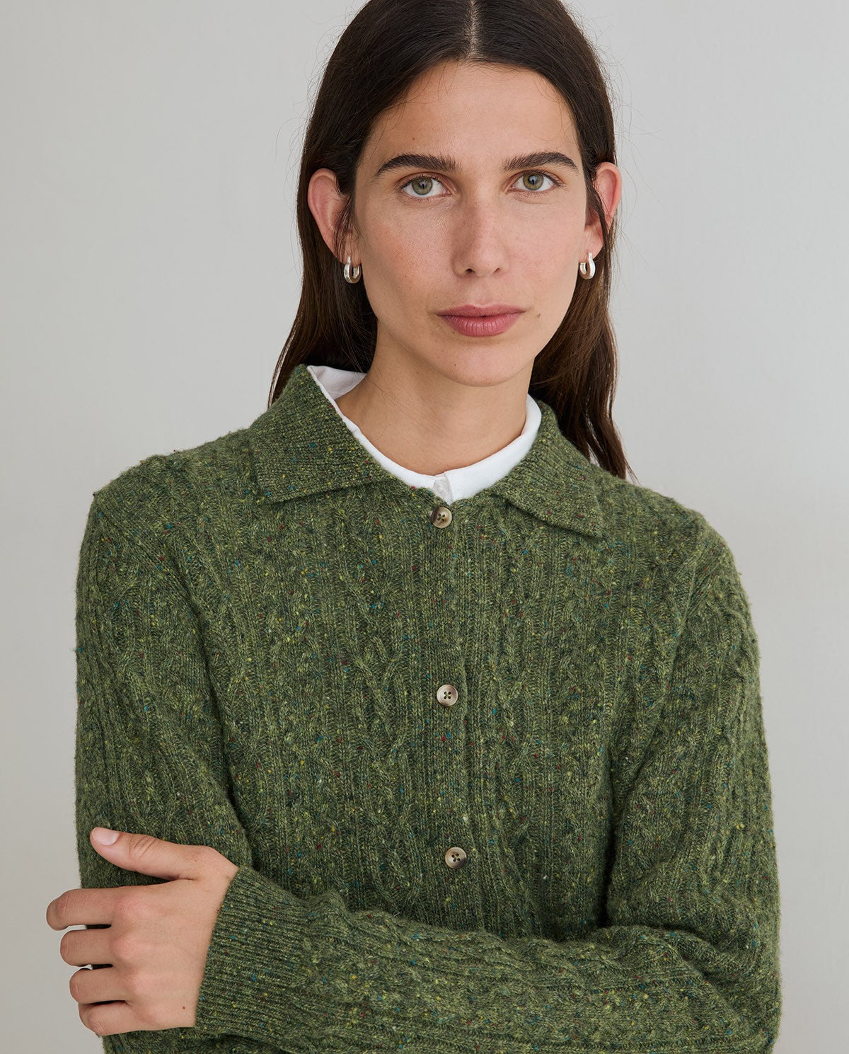 A woman wearing a textured olive green cable-knit button-up cardigan sweater over a white crewneck shirt, photographed against a neutral light gray background.