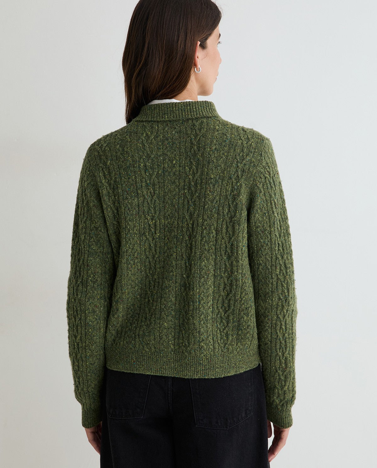 A woman viewed from the back wearing an olive green cable-knit sweater over a white collared shirt and black pants, against a plain light gray studio background.