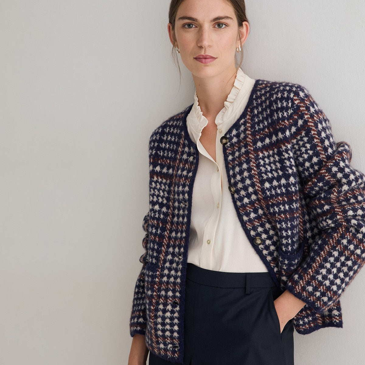 A woman models a navy blue and cream houndstooth tweed jacket over a cream ruffled blouse and navy trousers, standing against a neutral light gray wall.