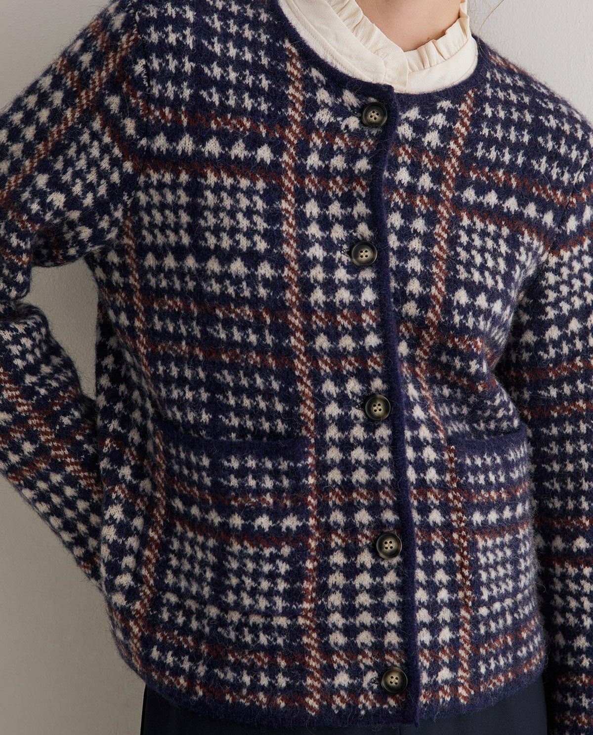 A close-up of a person wearing a textured knit cardigan with a navy, cream, and rust-brown houndstooth and windowpane pattern, layered over a cream ruffled blouse.