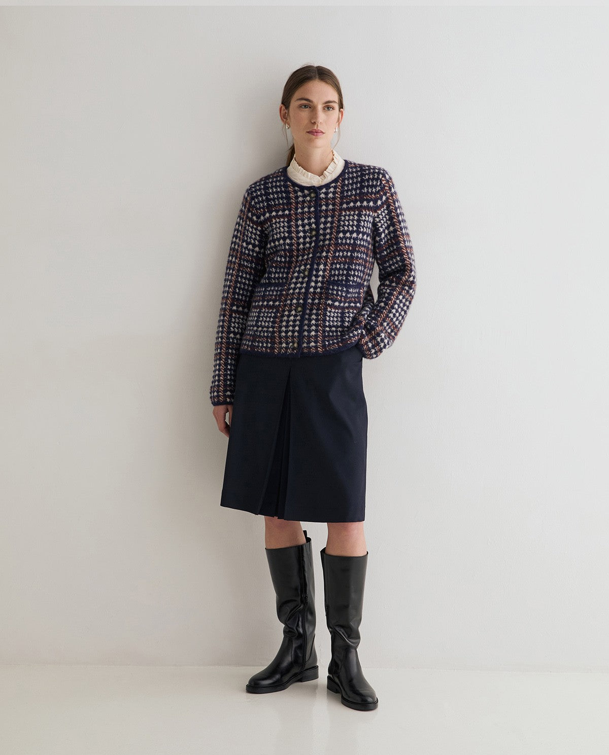 A model stands against a plain white studio wall wearing a navy, red, and beige tweed jacket over a cream collar, paired with a dark navy skirt and black knee-high boots.