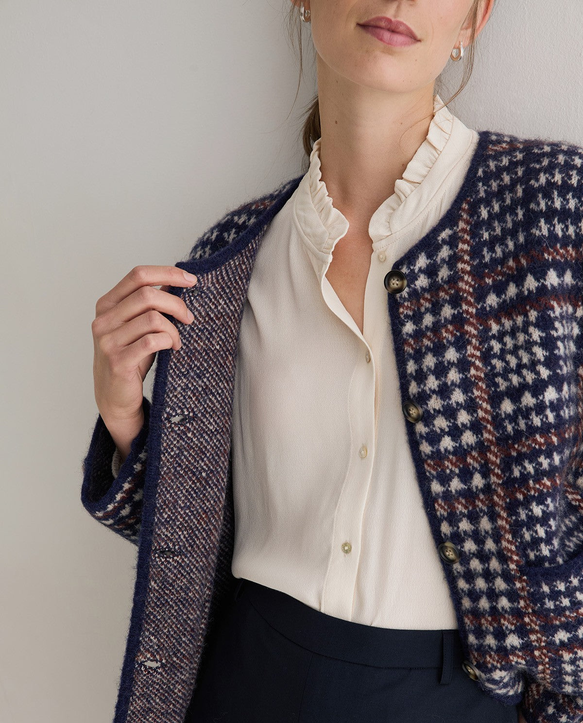 A woman wearing a navy blue houndstooth patterned cardigan over a cream ruffled blouse and navy trousers, holding the cardigan open against a plain light gray background.