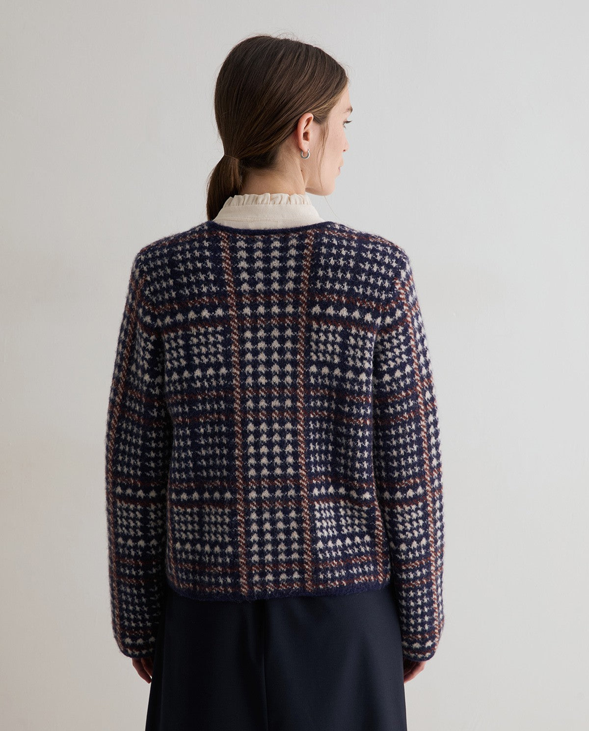 A woman viewed from the back wearing a dark navy and cream plaid knit sweater with a ruffled cream collar, paired with a dark skirt, against a plain light gray background.
