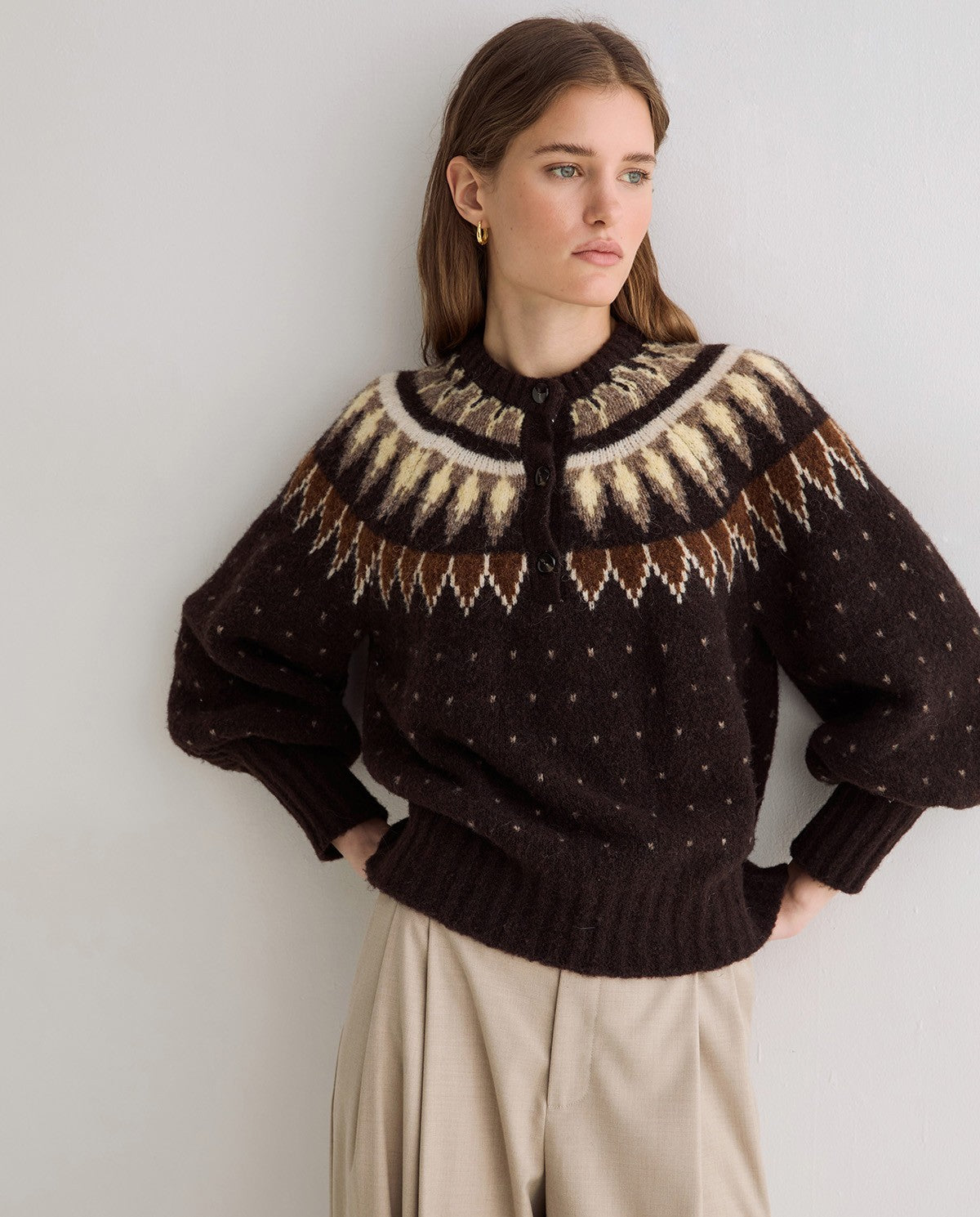 A woman wearing a dark brown knit sweater with a cream and brown Fair Isle yoke pattern, paired with beige trousers, standing against a light gray wall.