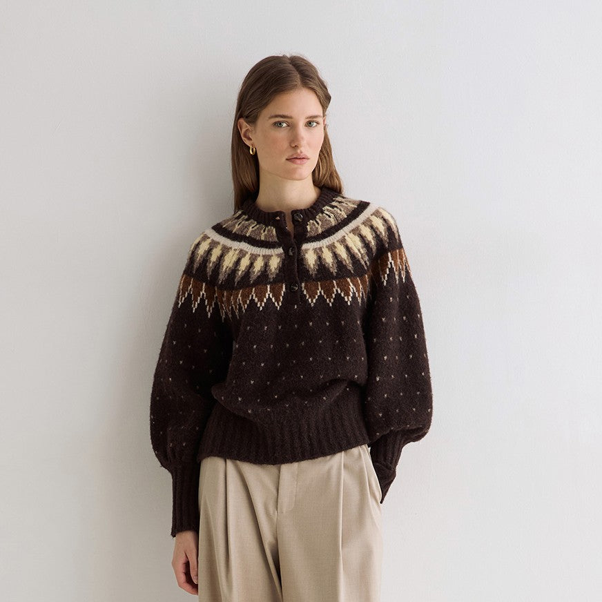 A woman stands against a plain white wall wearing a dark brown knit sweater with a cream and brown Fair Isle yoke pattern, paired with wide-leg beige trousers.