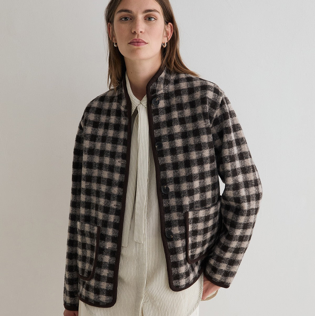 A woman modeling a black and beige checkered cropped jacket over a cream ribbed knit top and cream trousers, photographed against a neutral light gray studio background.