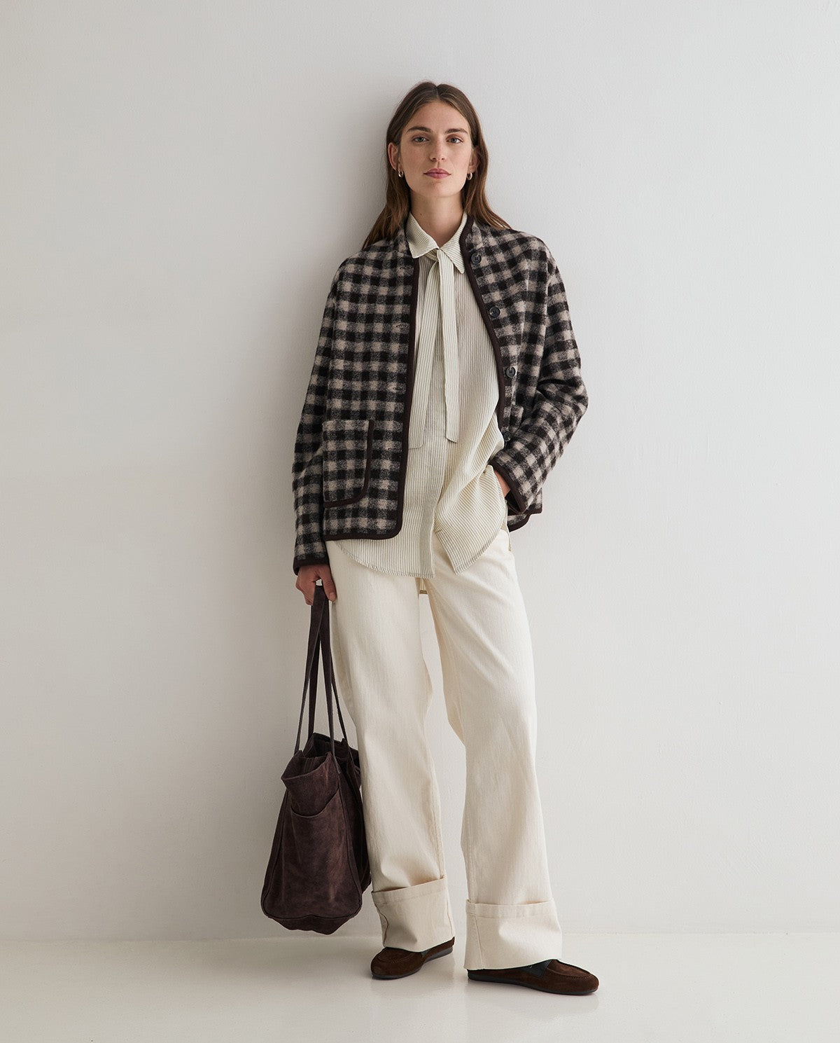 A woman stands against a plain light gray wall wearing a black and white plaid button-up jacket over a cream shirt and cream wide-leg trousers, holding a dark brown tote bag.