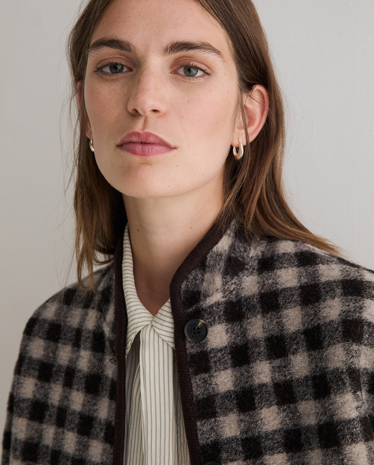 A close-up portrait of a woman wearing a black and beige checkered wool-blend jacket over a cream striped shirt, with small gold hoop earrings, against a plain light gray background.