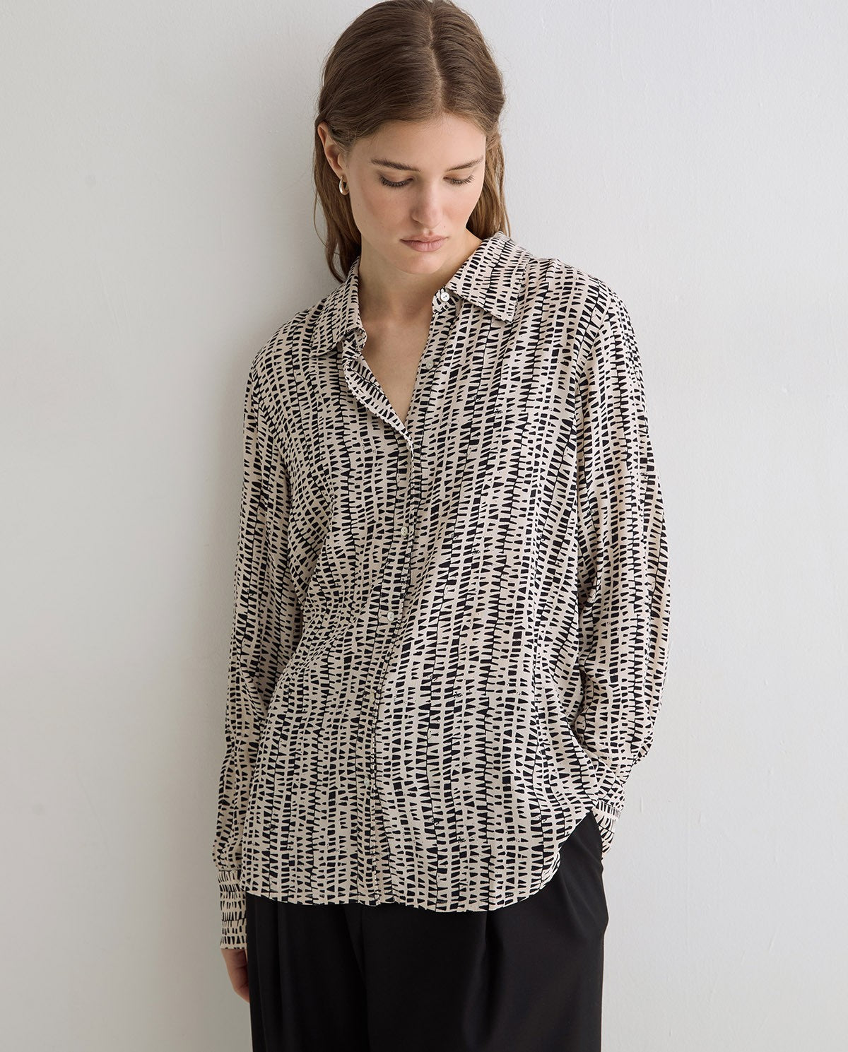 A woman wearing a black and white abstract patterned button-up shirt and black trousers, standing against a plain light gray wall.