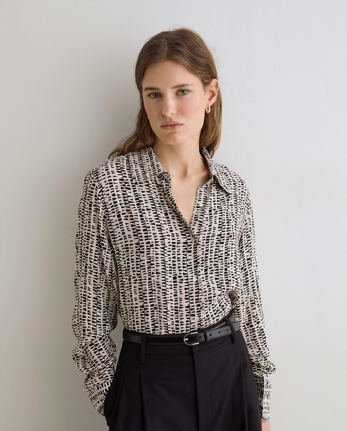 A woman wearing a black and white abstract patterned button-down shirt tucked into black trousers with a black belt, standing against a light gray wall.