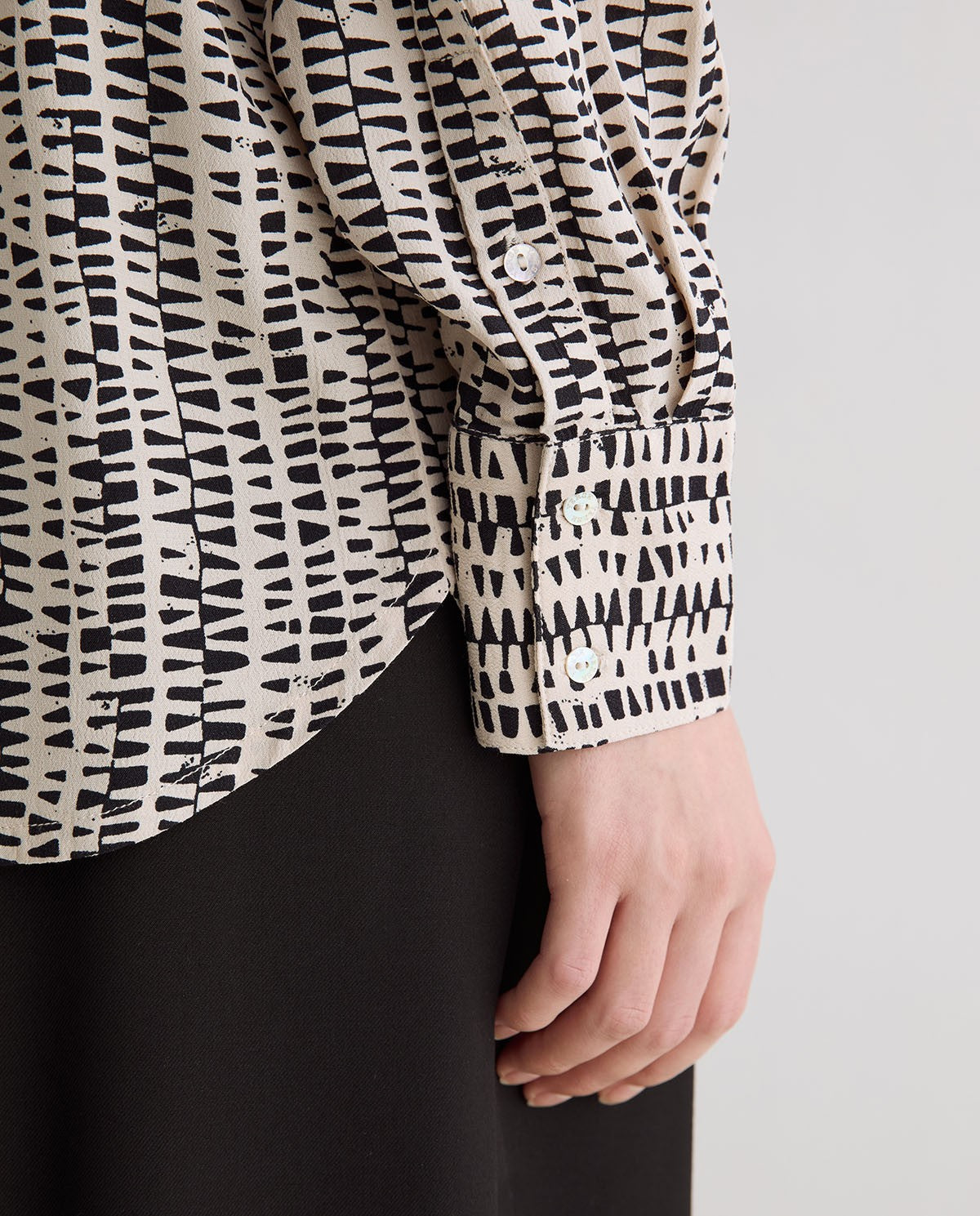 Close-up of a person wearing a long-sleeve blouse with a black and off-white abstract brushstroke pattern, paired with a black skirt or pants, against a plain light gray background.