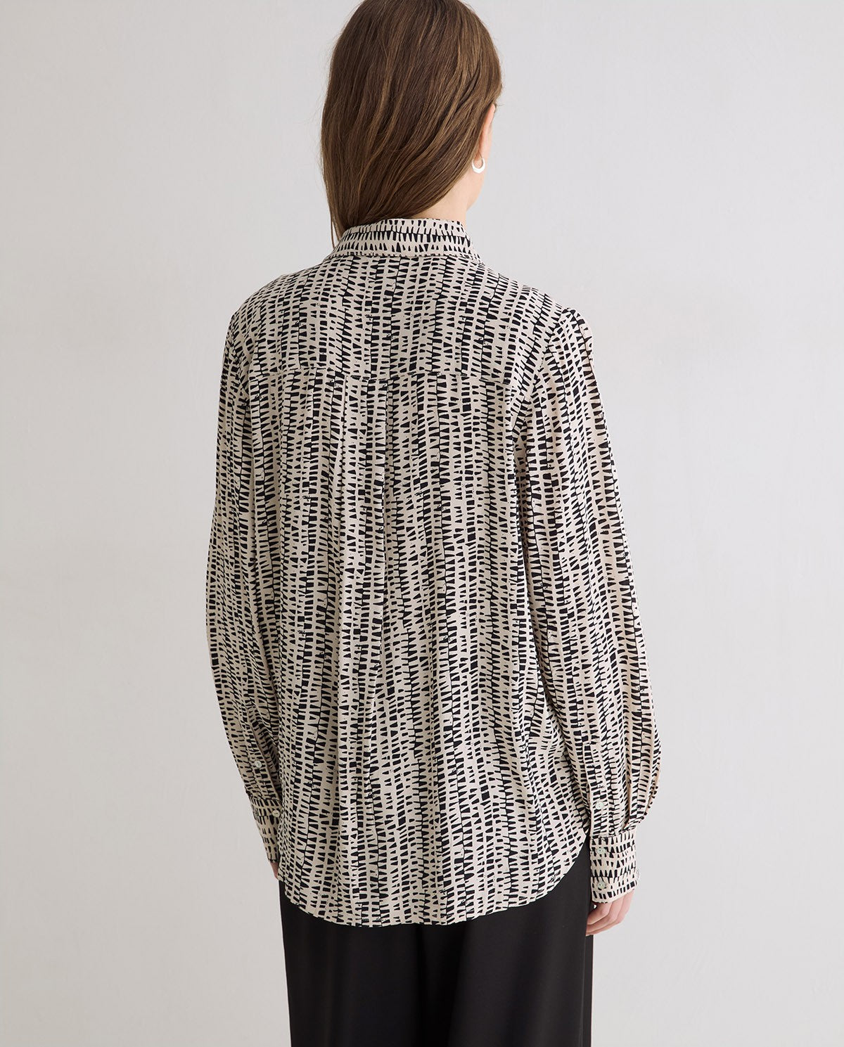 A woman viewed from the back wearing a black and white textured abstract patterned long-sleeve blouse and black trousers, set against a plain light gray studio background.