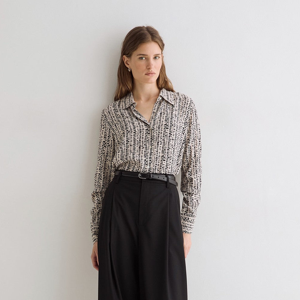 A woman stands against a plain light gray wall wearing a black-and-white patterned button-up shirt and high-waisted black wide-leg trousers with a black belt.