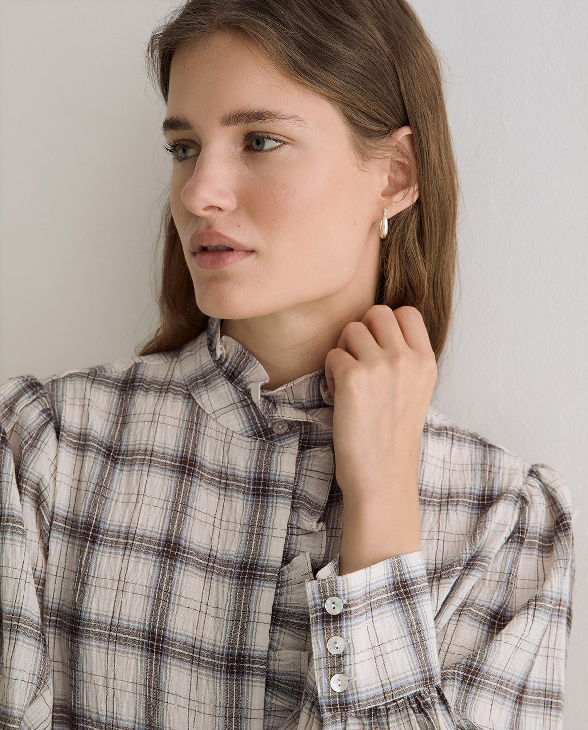 A woman wearing a beige and gray plaid button-up shirt, shown from the chest up against a plain light gray wall.