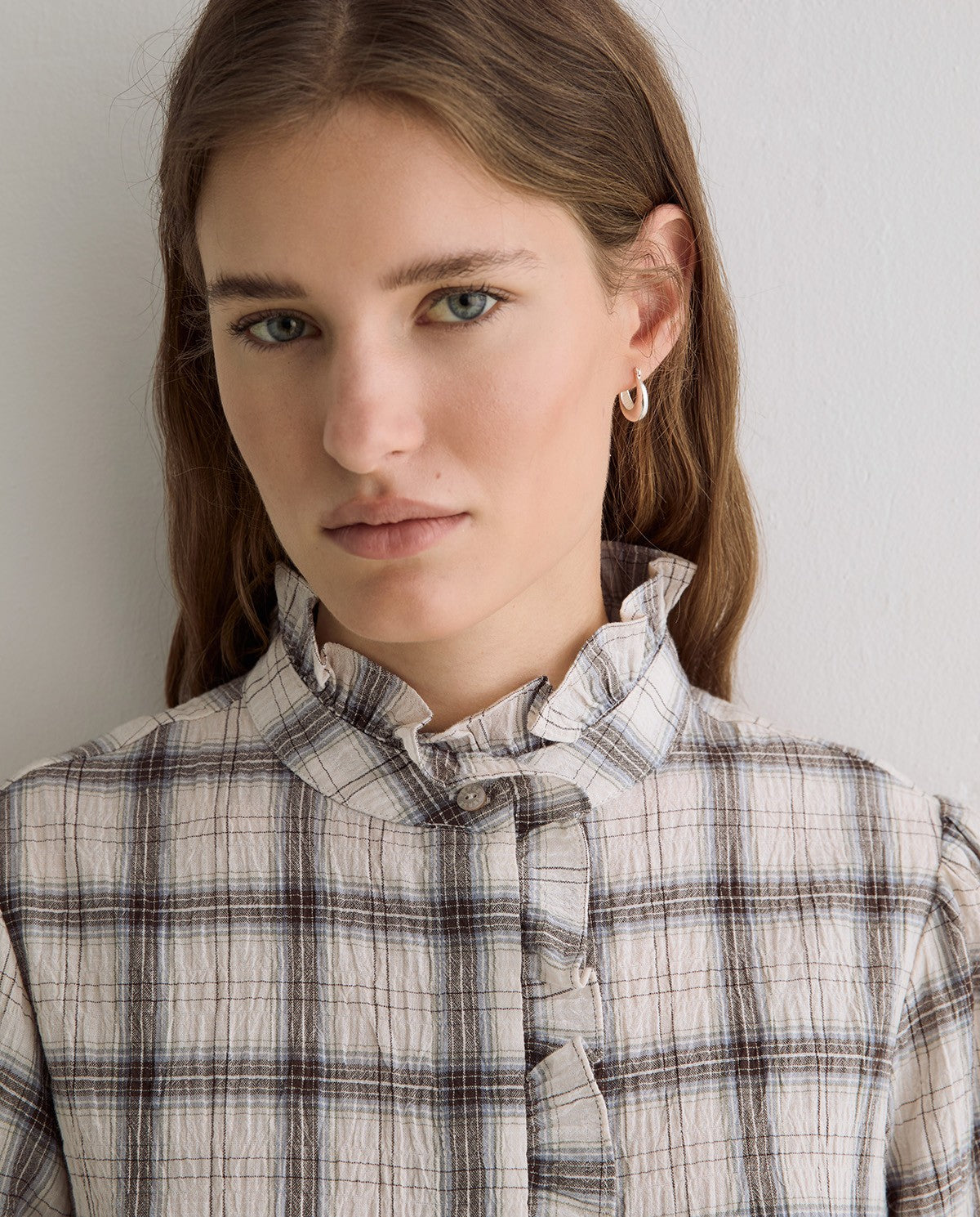 A close-up shot of a woman wearing a beige and brown plaid shirt with a ruffled collar, set against a plain light gray wall.