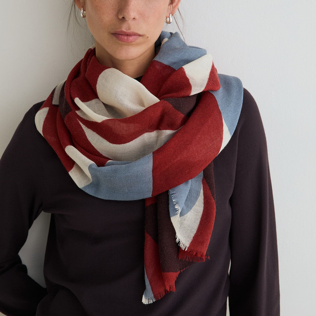 A woman wearing a large woven scarf with a block pattern in burgundy red, light blue, and cream, styled over a dark plum top against a plain light gray wall.