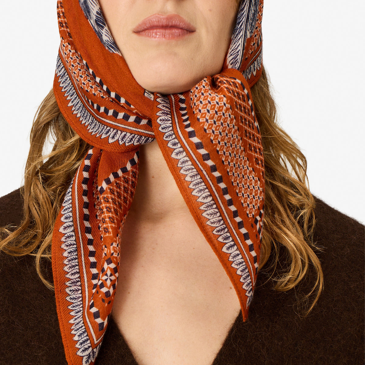 A close-up of a woman wearing a patterned orange scarf tied around her neck, paired with a brown knit sweater against a plain light background.