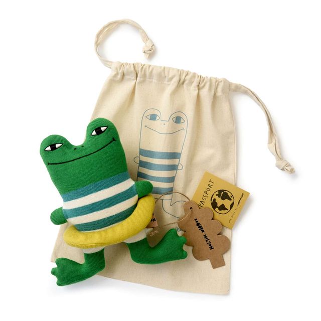 A green plush frog toy with a blue and white striped shirt and yellow belly, posed in front of a cream canvas drawstring bag with a frog illustration. A small brown tag and yellow passport-style tag are attached, all on a white background.