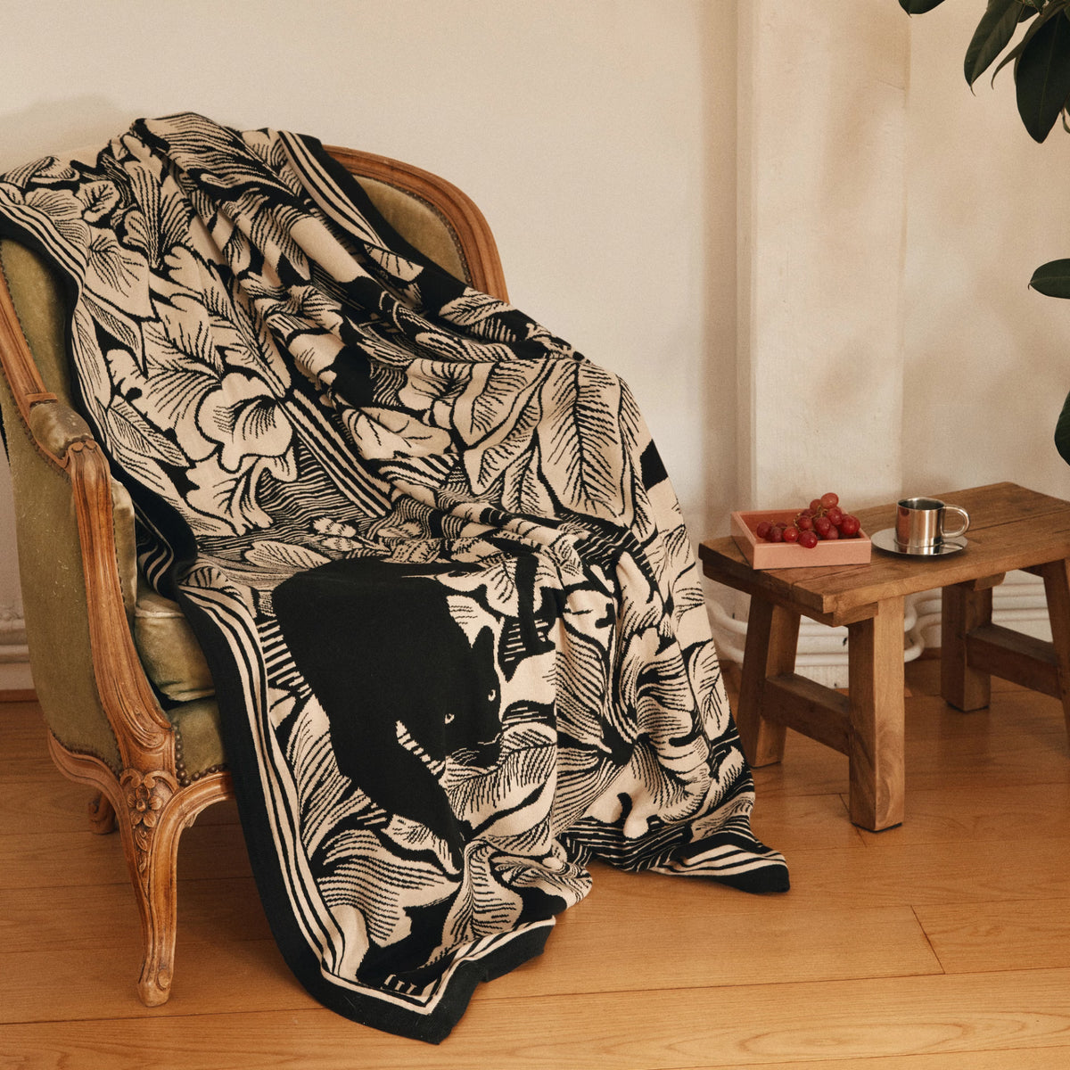 A black and cream botanical throw blanket with a large black cat motif draped over an antique green armchair in a warm, rustic room next to a wooden stool.