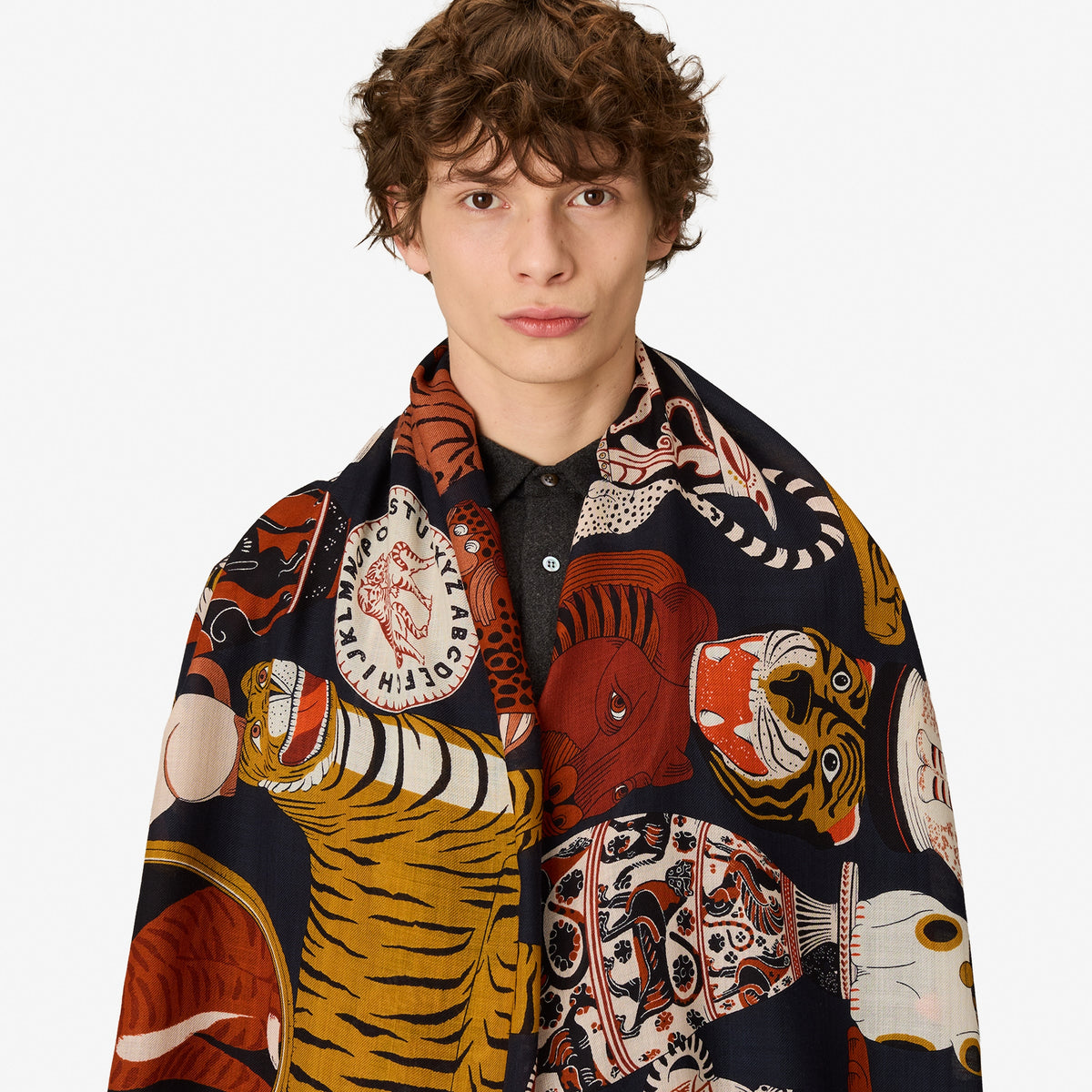 A model with curly brown hair wearing a black collared shirt and draped over his shoulders is a large, colorful scarf featuring a classic multi-species illustration print of tigers, snakes, and fish on a dark navy background.