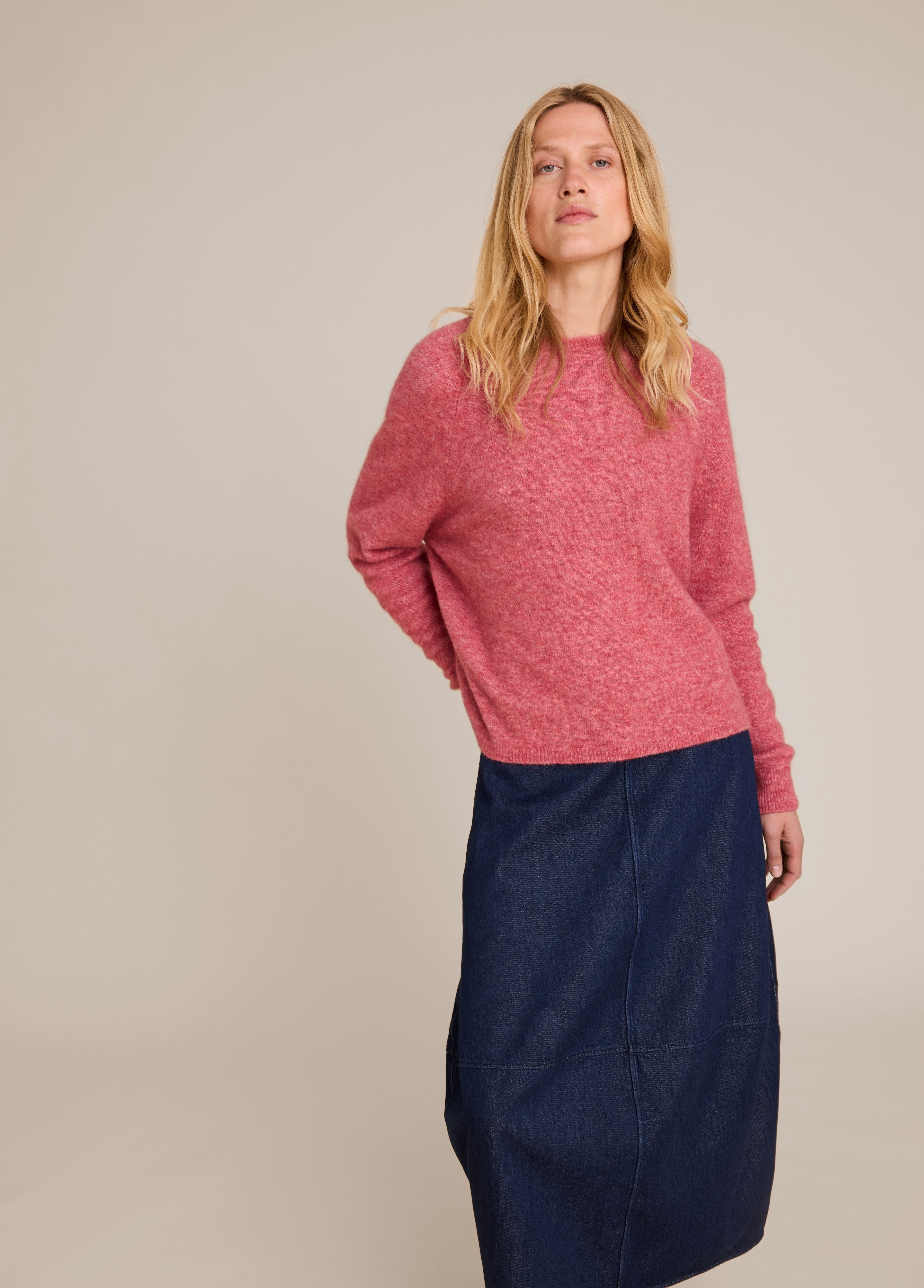A woman modeling a heathered pink knit sweater paired with a dark navy blue denim skirt, photographed against a solid light beige studio background.