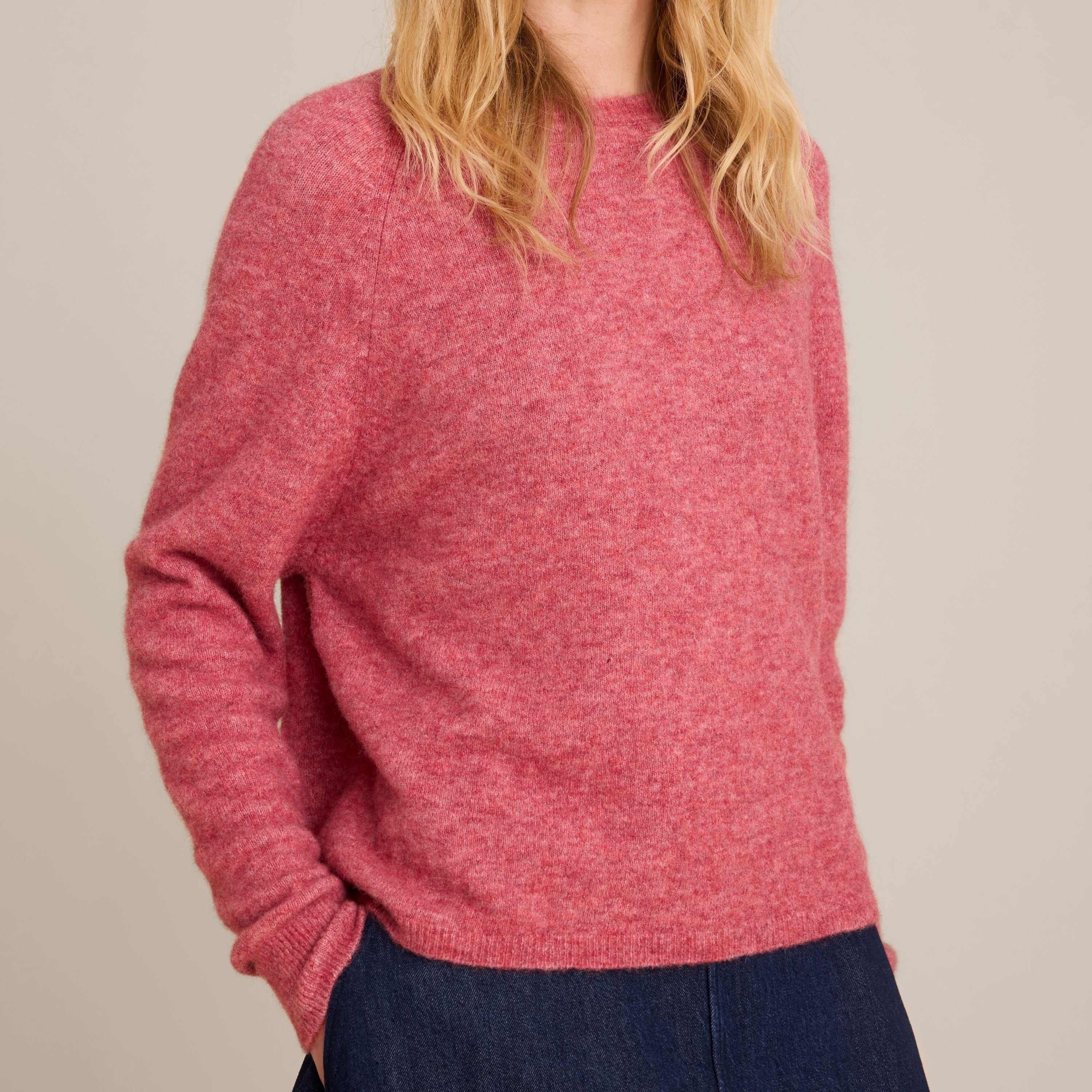 A woman with blonde hair wearing a heathered dusty rose pink knit sweater and dark navy pants, standing against a plain light beige studio background.
