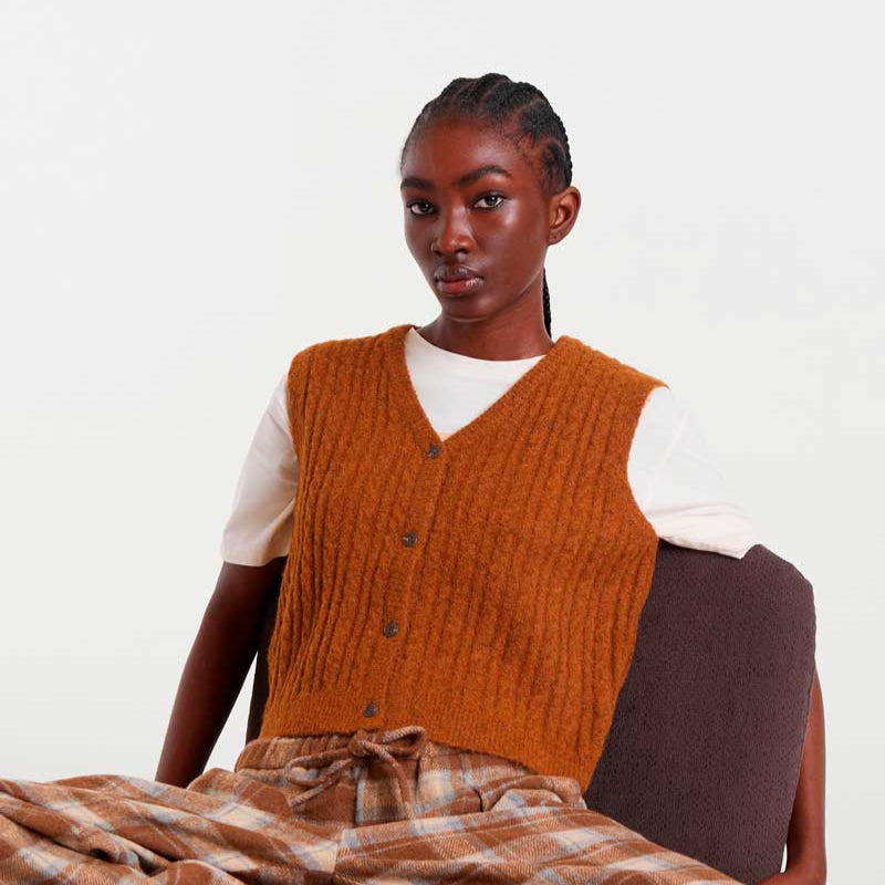 Person wearing a brown knitted vest and plaid pants sitting on a chair.