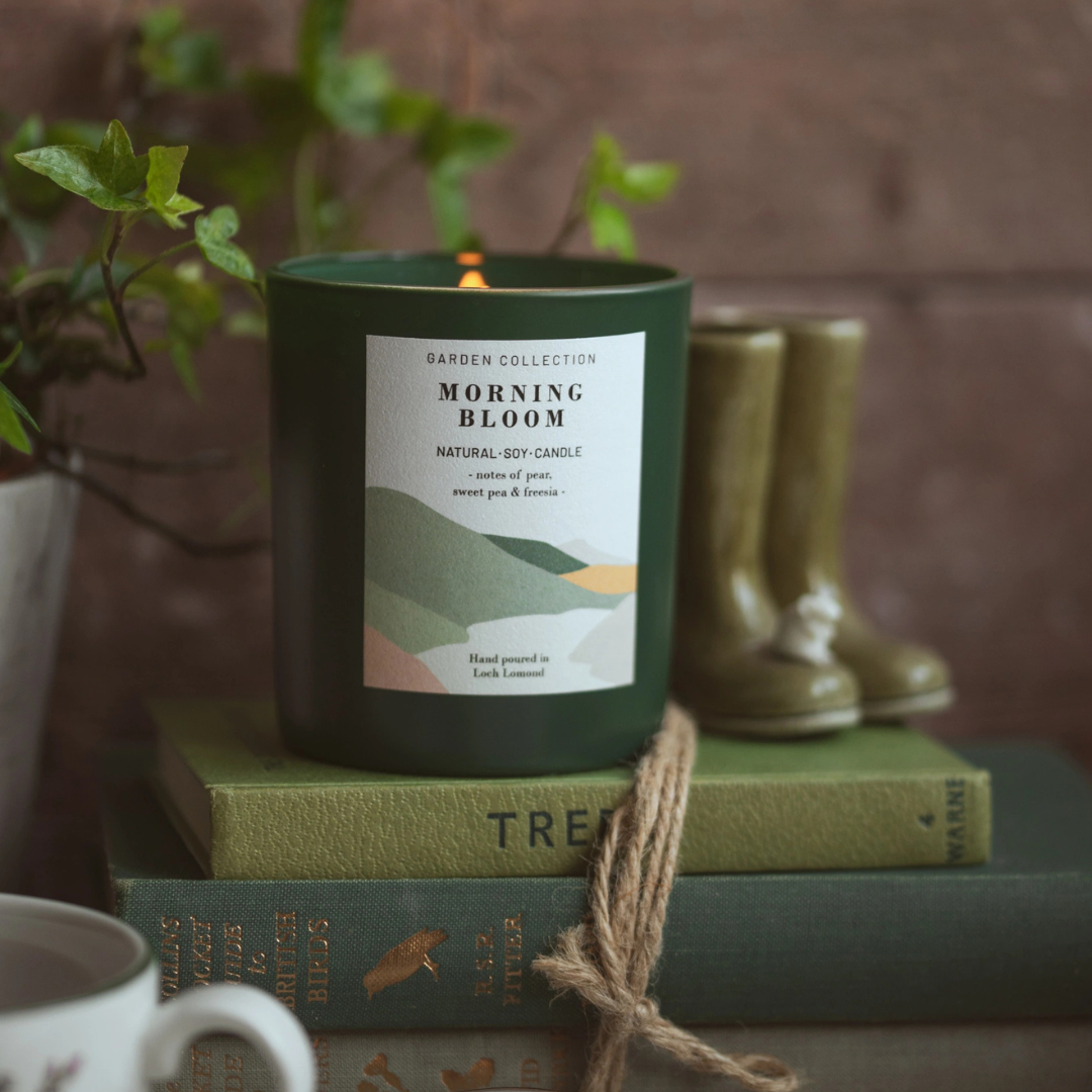 A dark green jar candle labeled 'GARDEN BLOOM' sits lit on stacked vintage books, with a small plant and green rain boots in the background against a rustic wood wall.