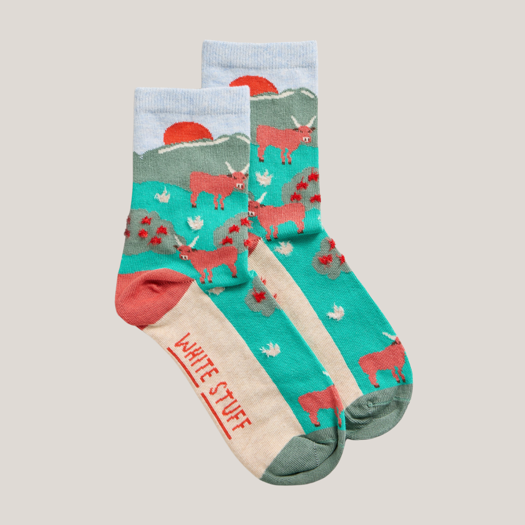 Pair of ankle socks featuring a teal and cream design with a repeating pattern of red Highland cows, a sunset, and small