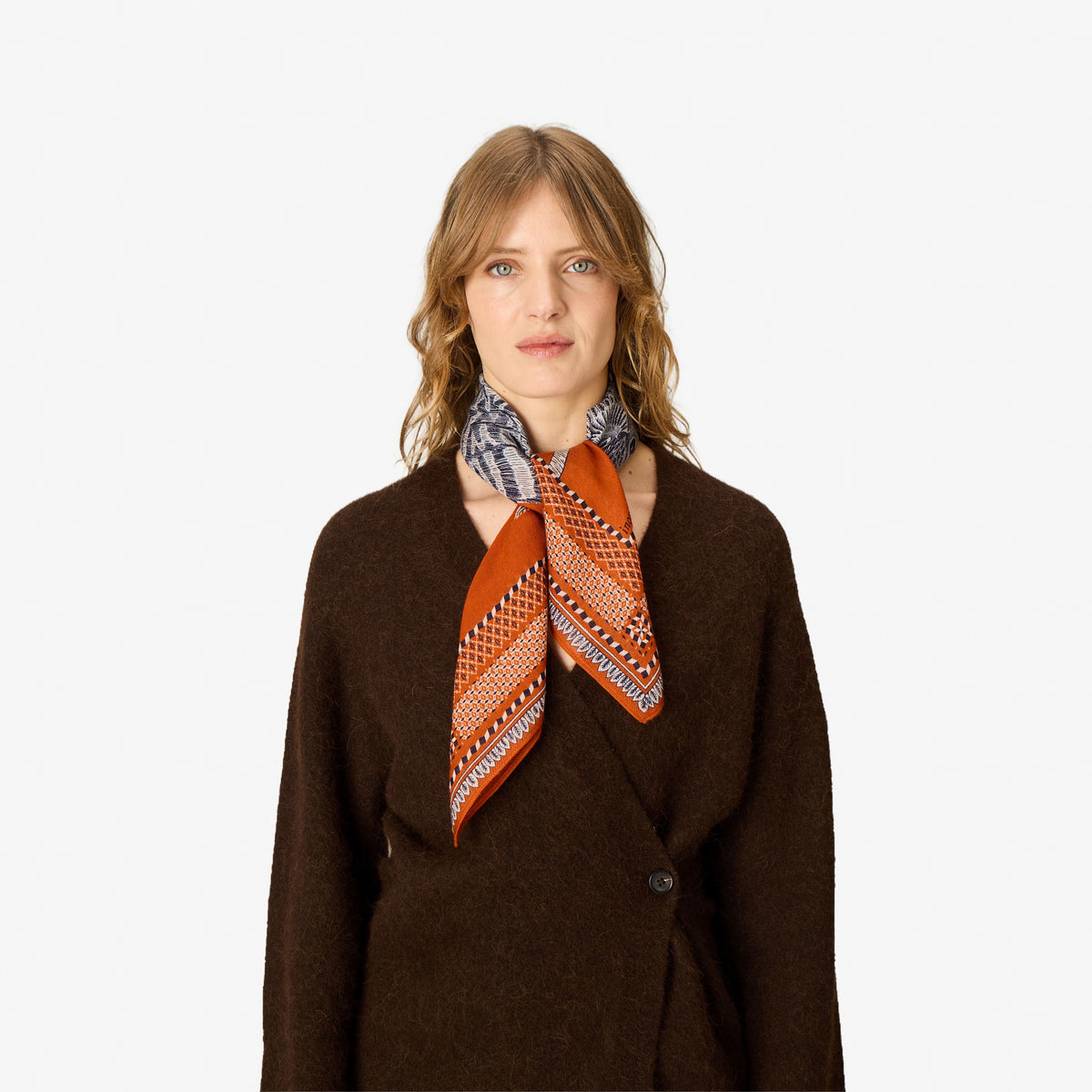 A woman wearing a brown cardigan and a patterned orange scarf, photographed against a plain white studio background.