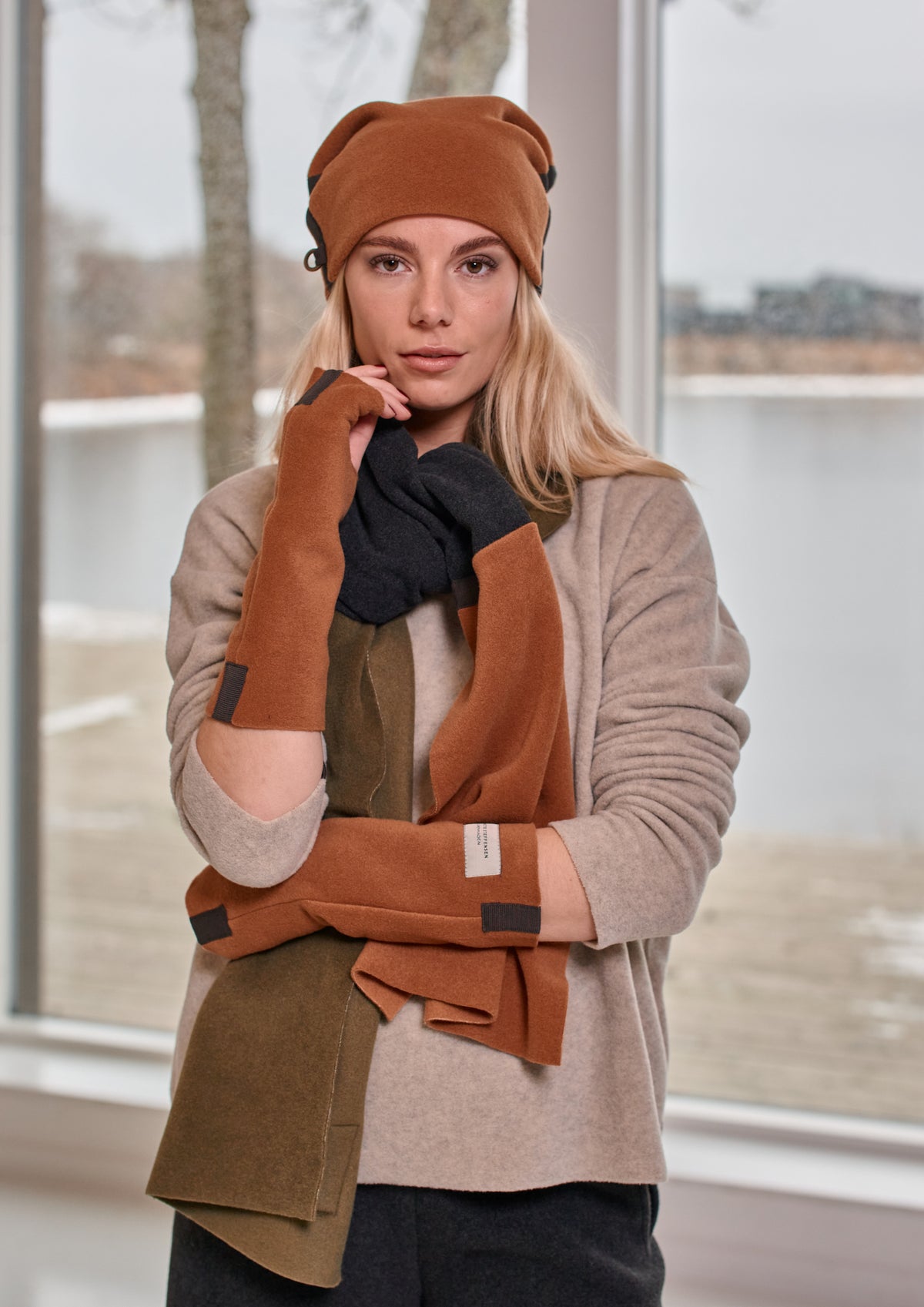 A woman modeling a coordinated autumn outerwear set consisting of a camel brown beanie, a multi-layered scarf in camel brown, olive green, and black, and a beige knit cardigan, posed in front of a large window.