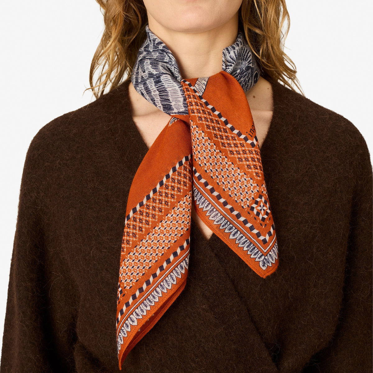 A close-up of a model wearing a brown knit sweater and a patterned silk scarf tied around the neck. The scarf features a rust-orange base with geometric diamond and lattice patterns in white, black, and gray.