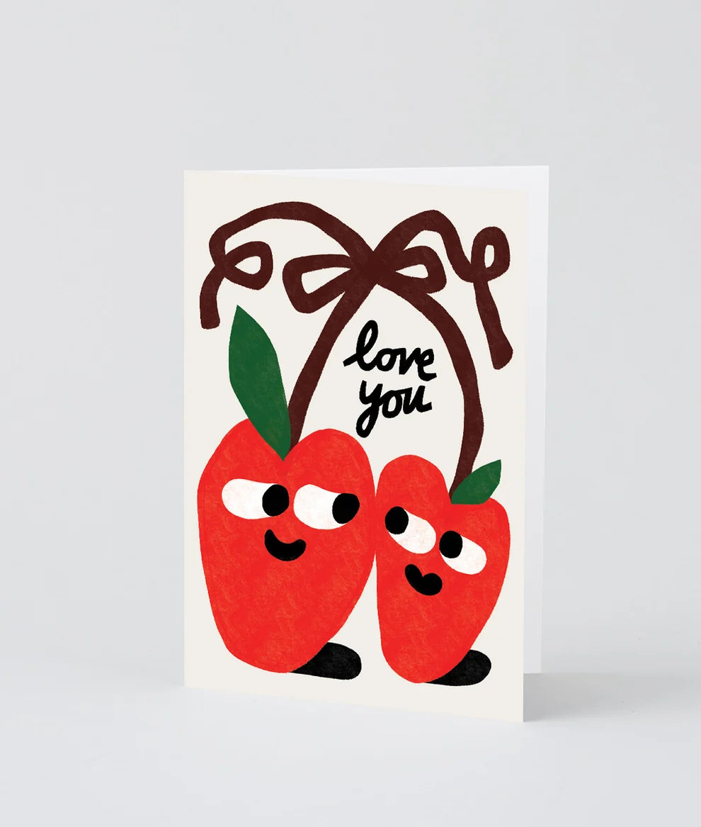 A vertical greeting card featuring two cute red apple characters with green leaves and black feet, tied under a large brown bow with the words 'love you' written in black script. The card is photographed standing upright on a light gray background.