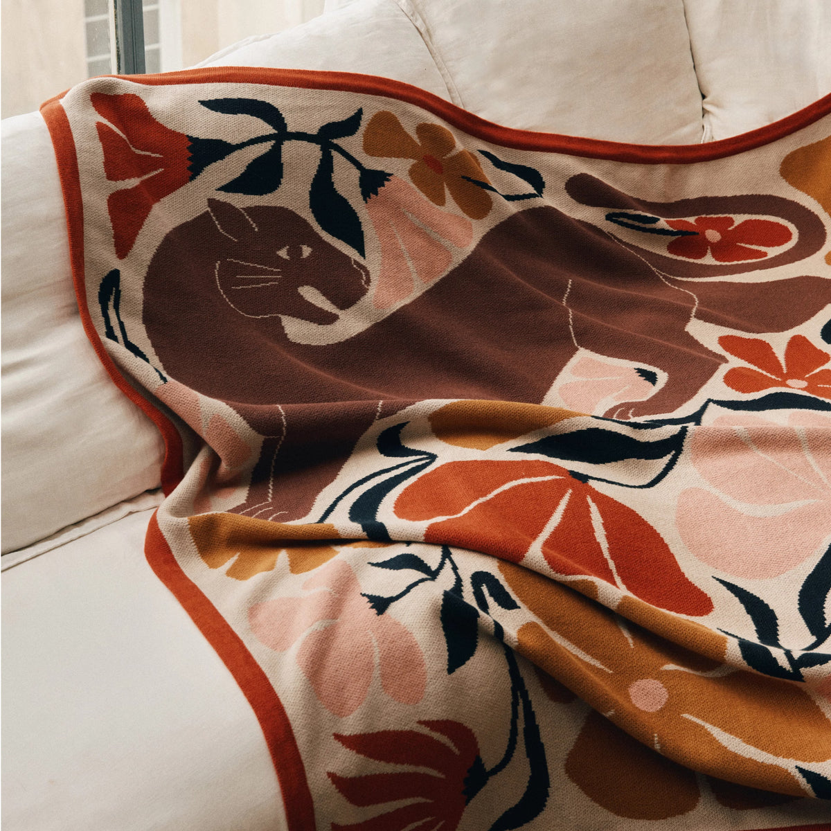 A cozy throw blanket draped over a cream-colored sofa, featuring a stylized brown cat and floral motifs in rust orange, peach, navy, and beige.