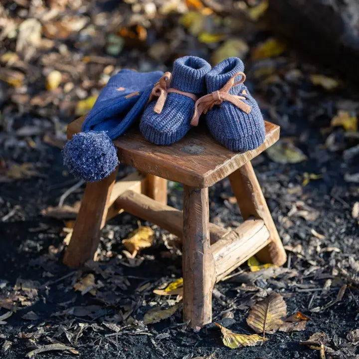 Pair of dark blue knitted baby booties with tan laces, and matching knitted hat with pompom, on a small wooden stool.